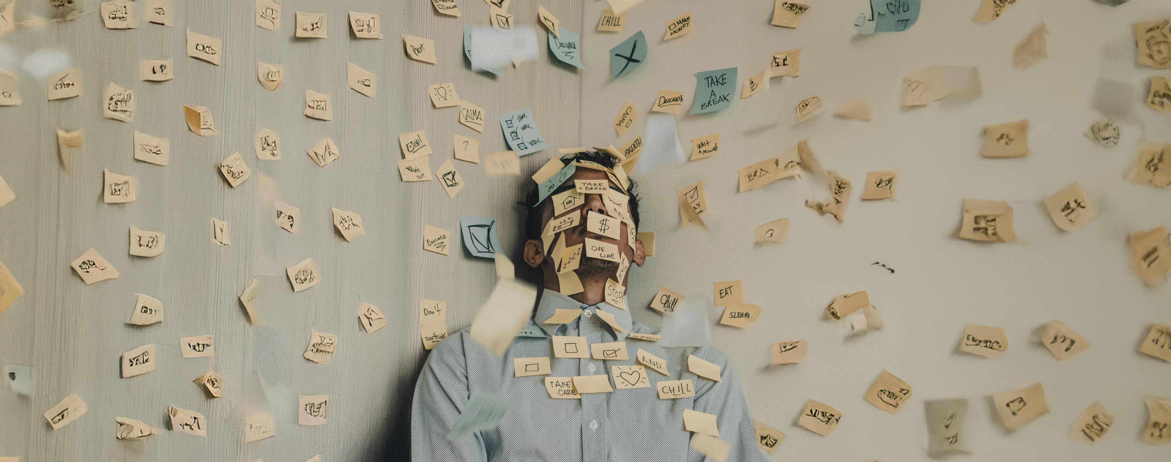 A man sits overwhelmed and covered in sticky notes, with hundreds more stuck to the wall behind him and floating in the air around him. The notes contain scribbles and reminders like "Take a break," "Chill," and "Eat," capturing a sense of burnout and mental overload.